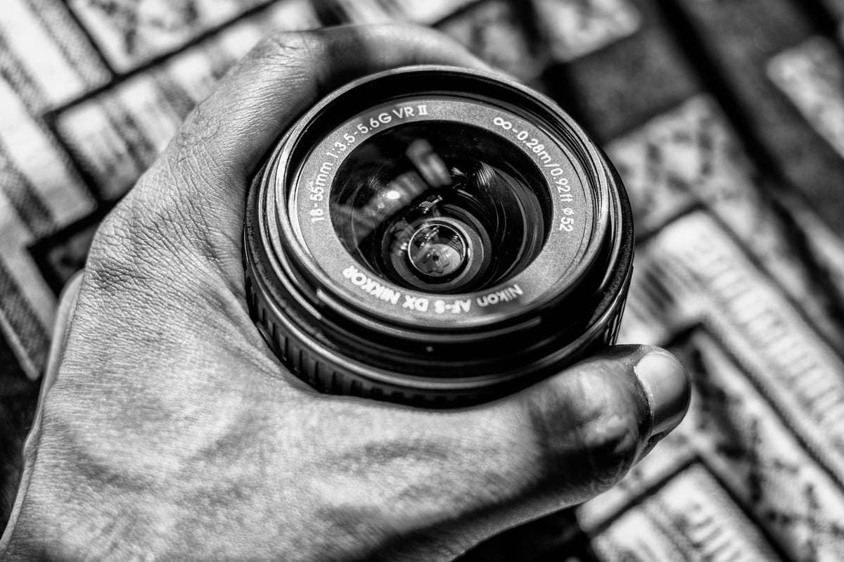 A black-and-white close-up of a camera lens held in a hand
